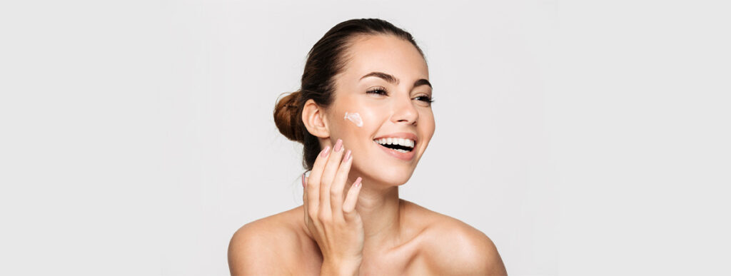A youthful woman applying sunscreen in her cheeks and smiling.